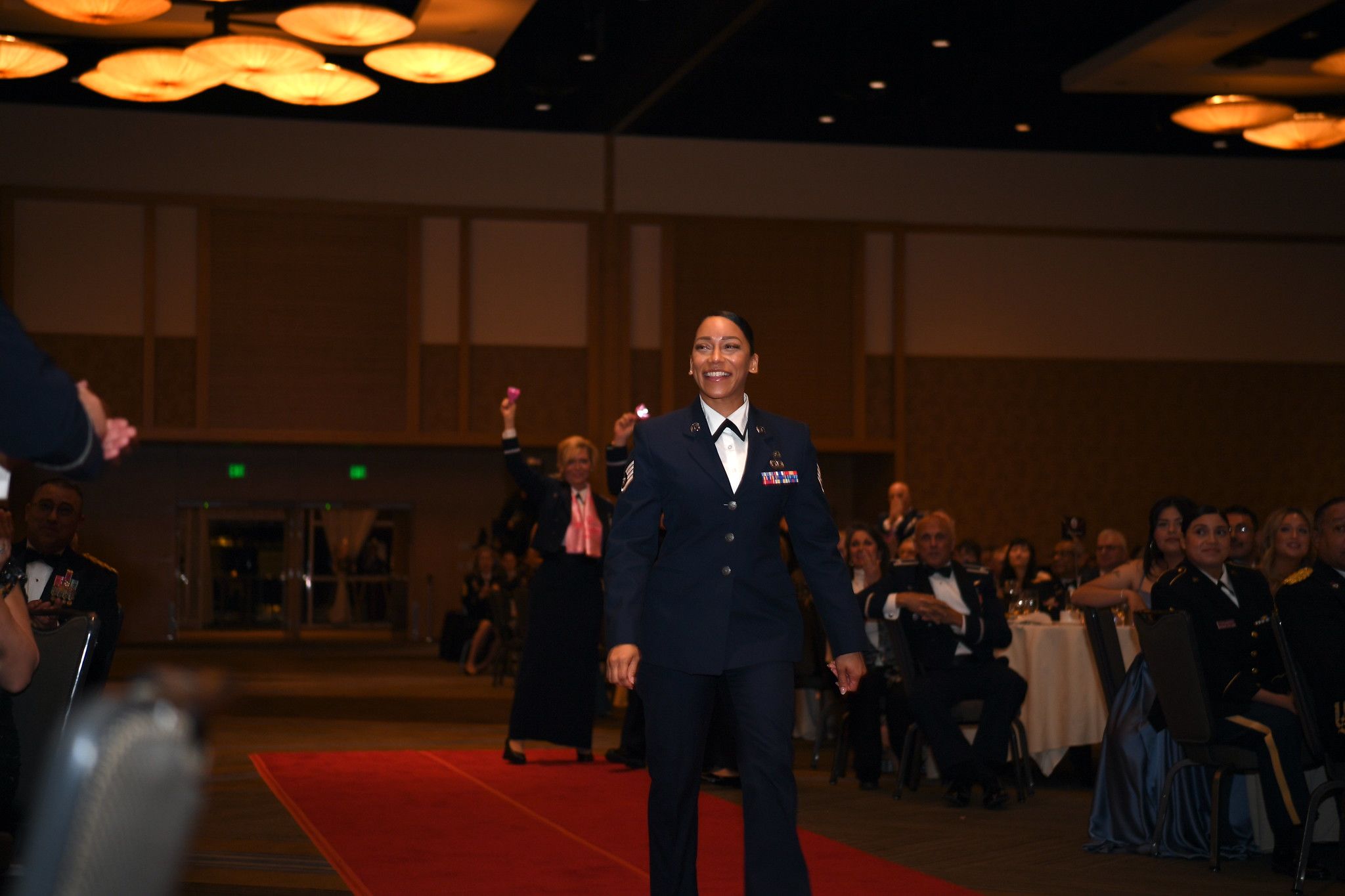 A woman in Air Force dress uniform smiles as she walks down a red carpet.