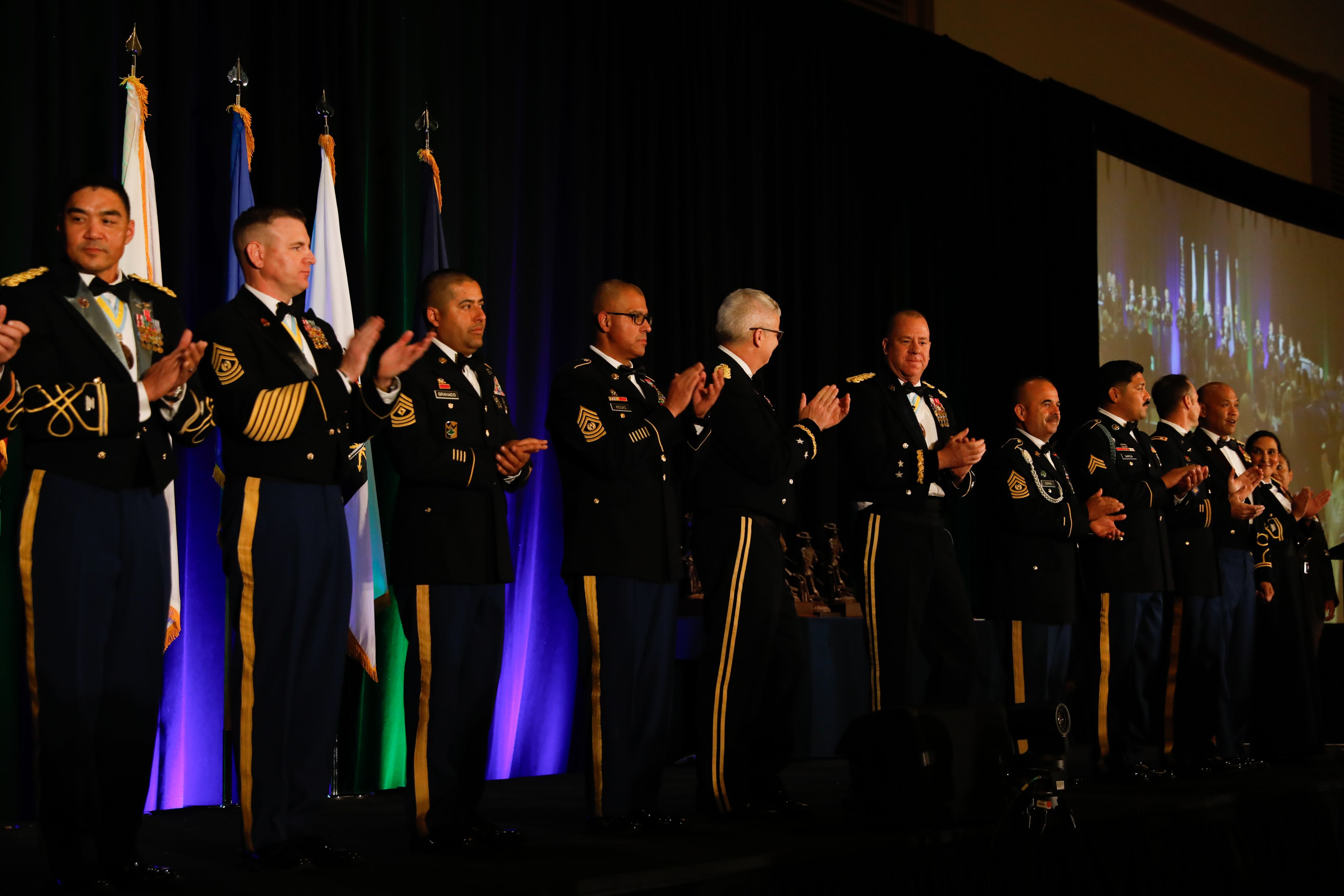 Soldiers in dress uniform stand on a stage and clap.
