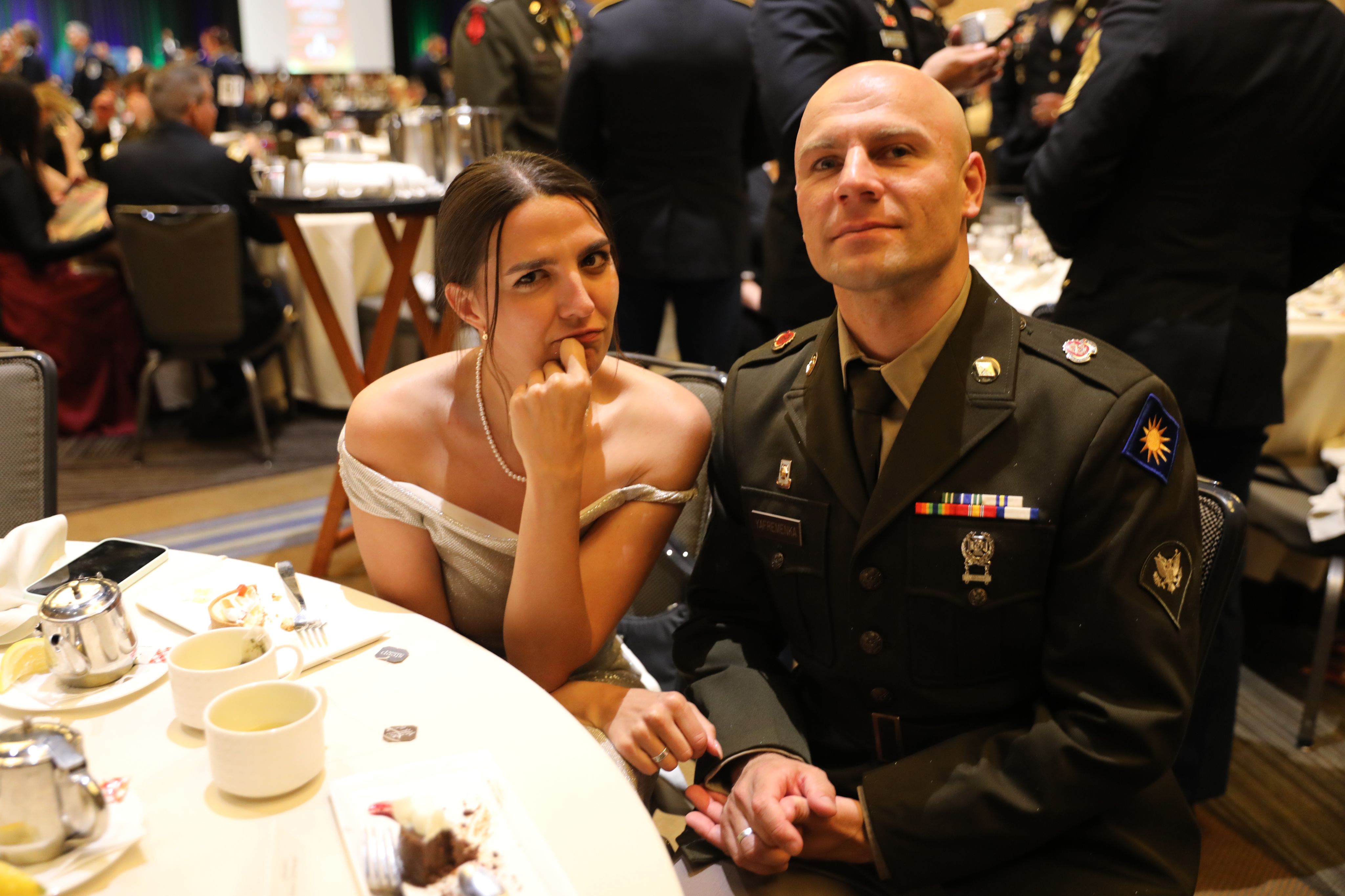 A Soldier in green dress uniform poses while seated at a banquet table next to a woman in a white evening gown.