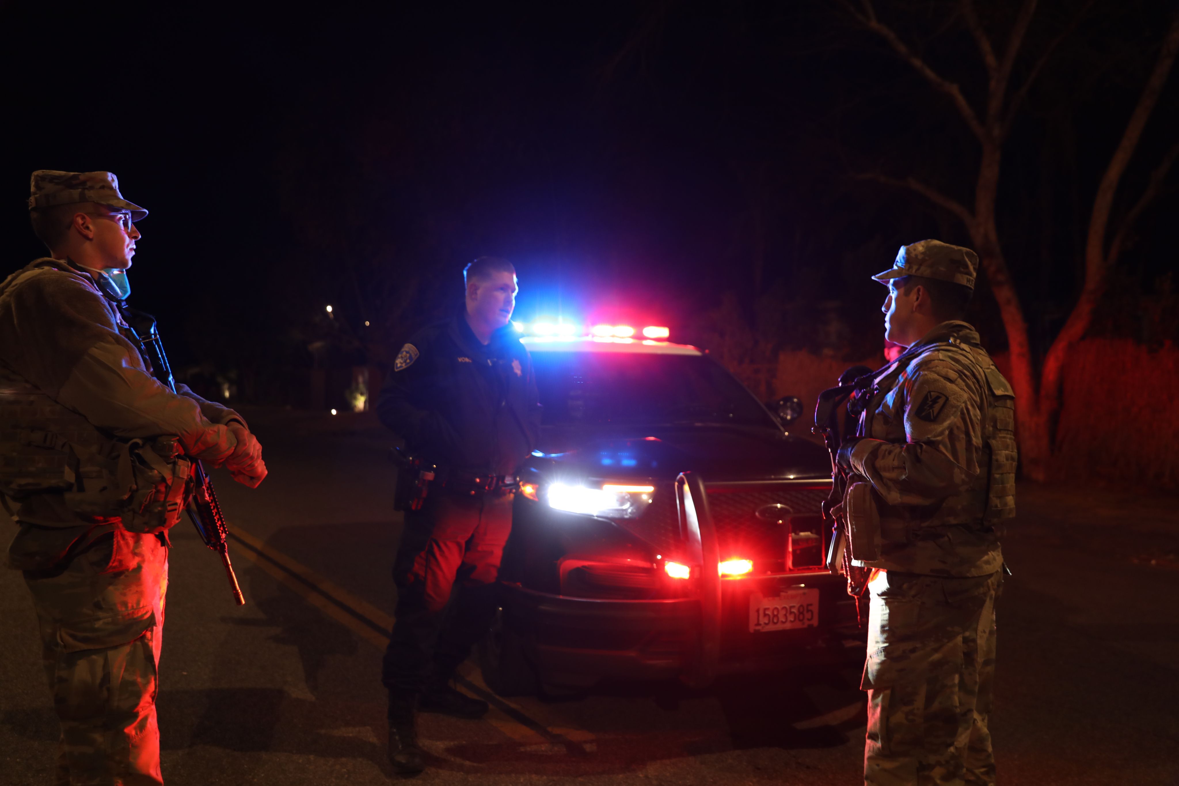 Two Soldiers and a police officer stand in front of a police car with its lights on.