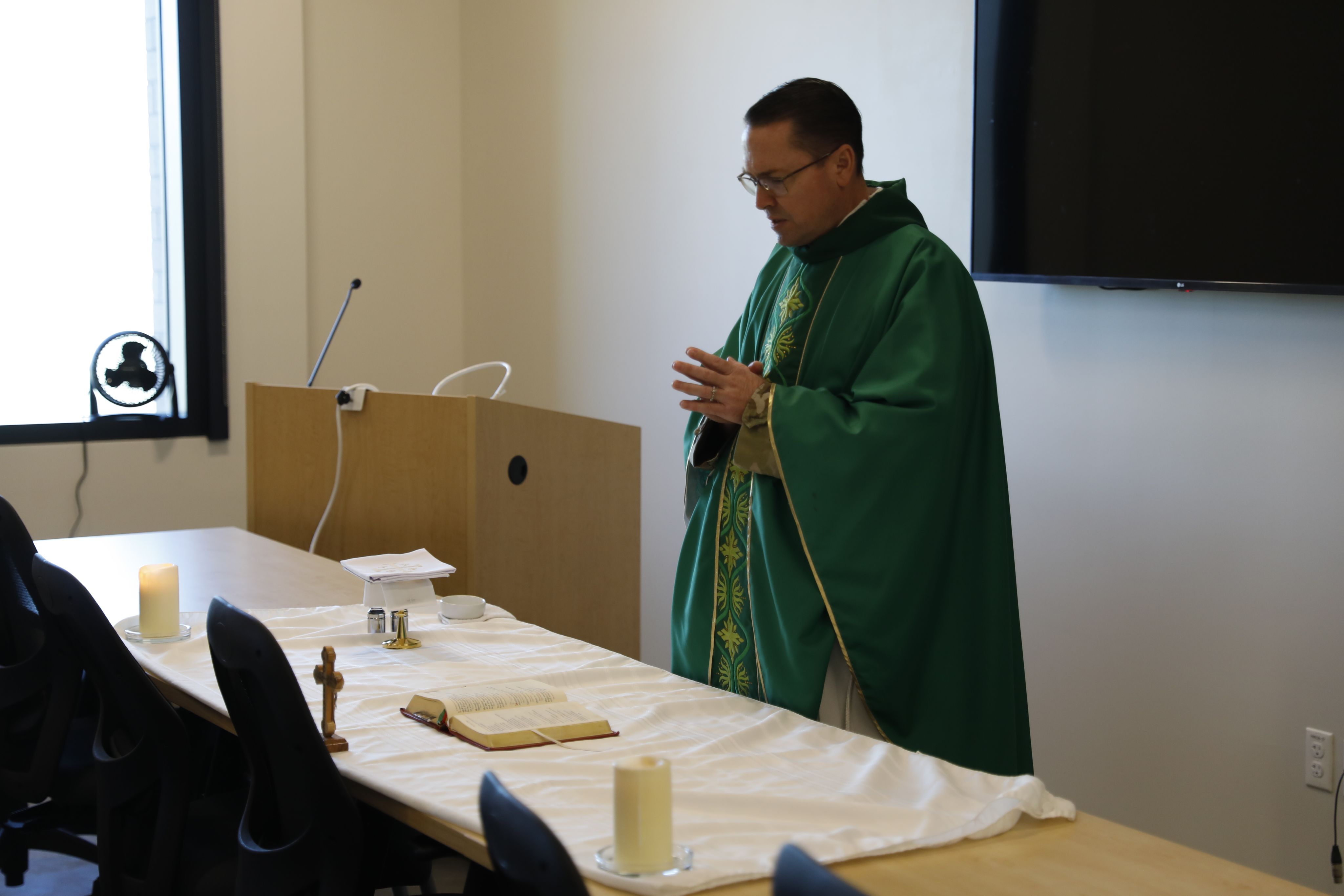 A chaplain stands behind a table with a missal, candles, a paten, a host, a corporal, a pall, a chalice, water, wine, an altar cloth, and a crucifix
