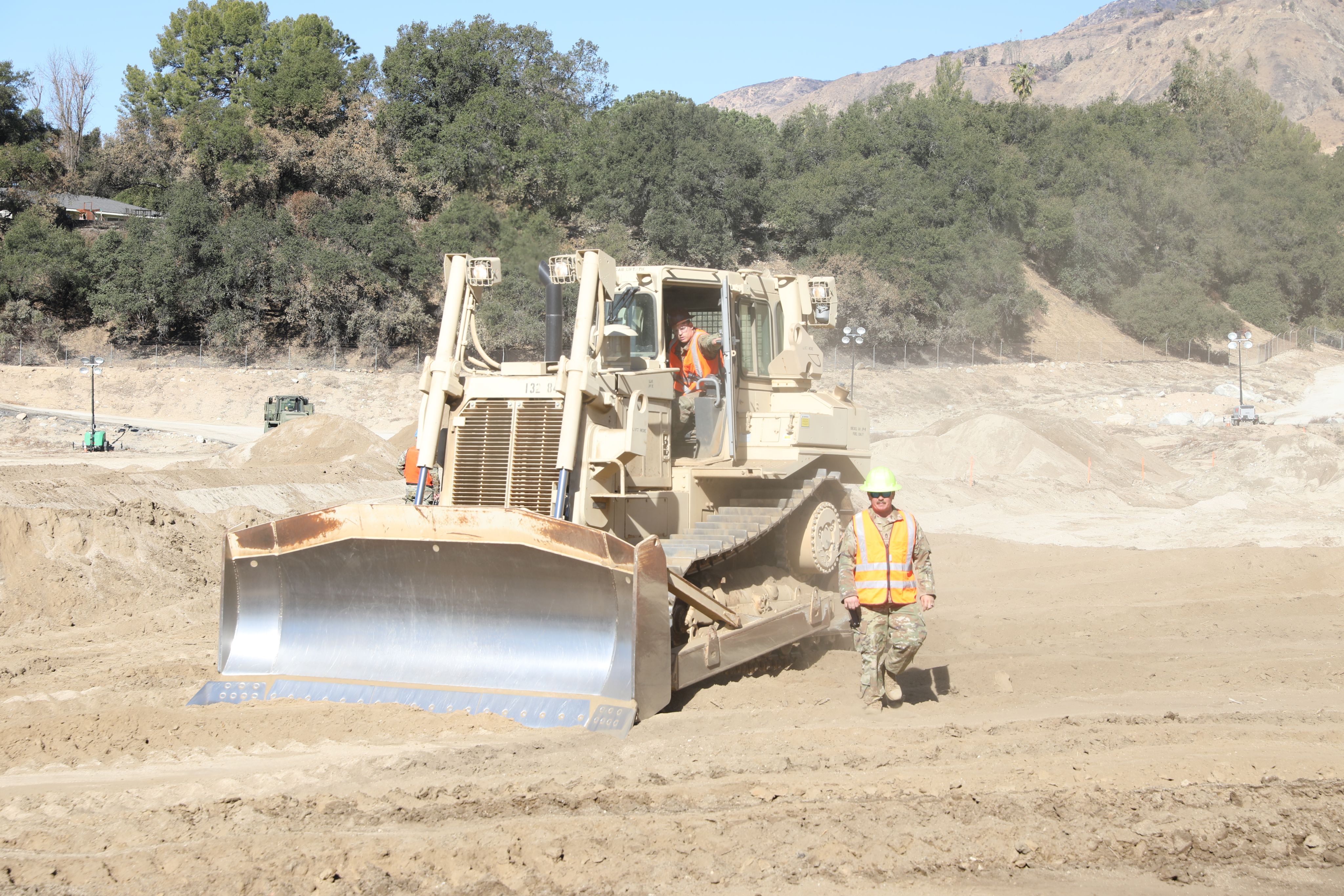 Two Soldiers in high visibility vests take a break from operating a tan bulldozer that has been pushing dirt in a debris basin.