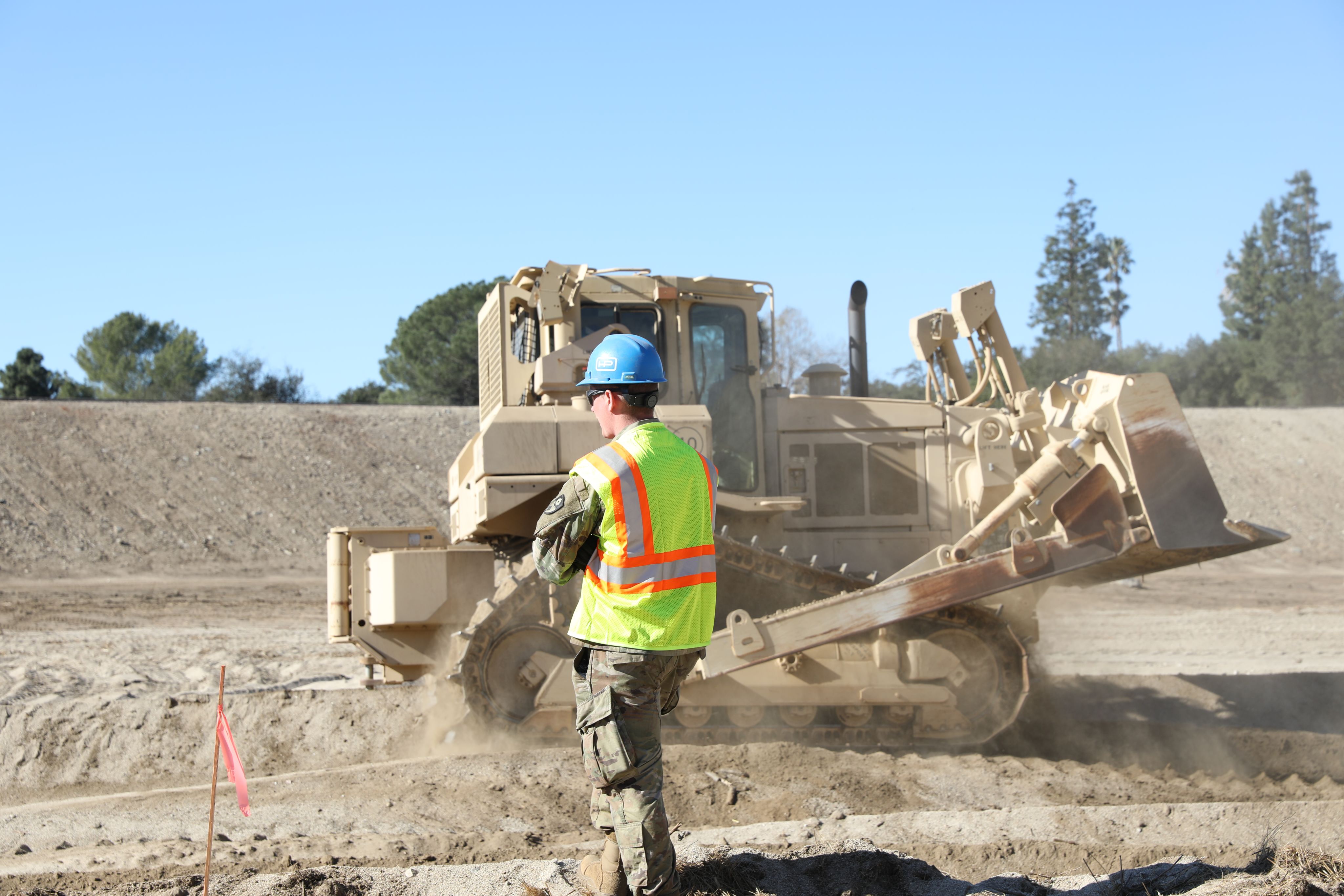 Soldier drives tan colored bulldozer in a dirt basin. with another soldier in high visibility vest and blue hard hat supervising the operation.