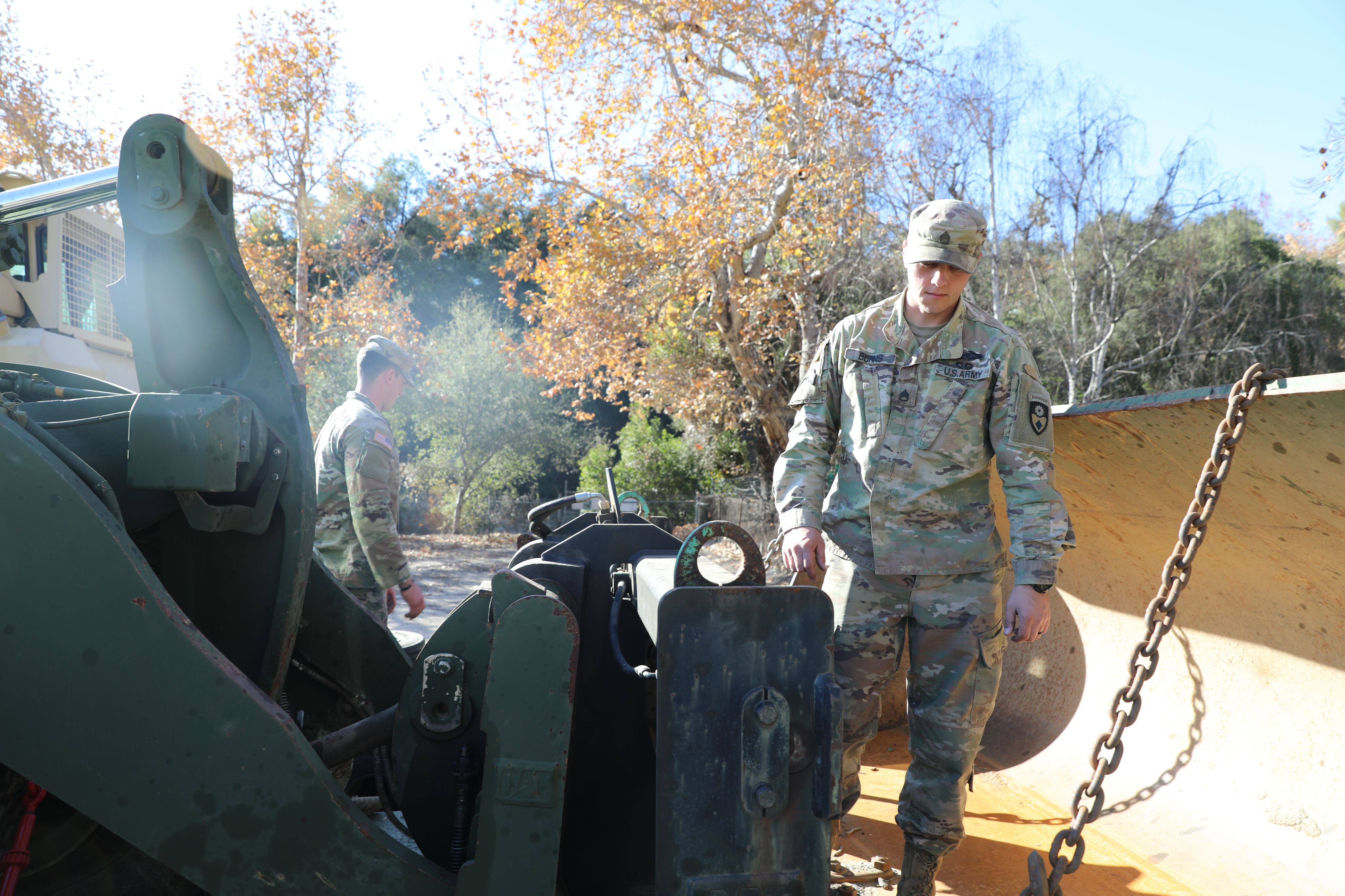 Two Soldiers stand in front of a green excavator in a wooded area.