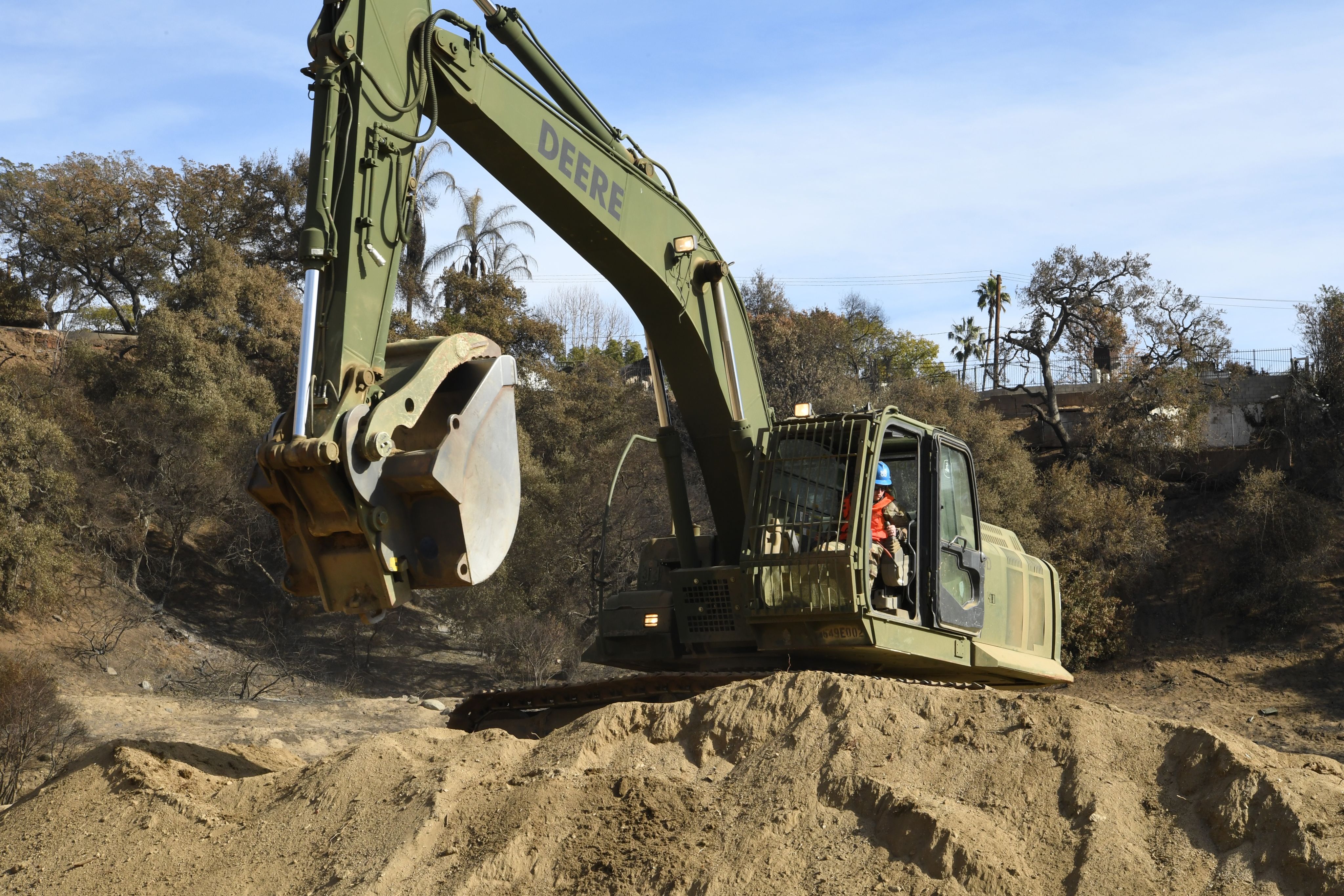A Soldier sits inside a green excavator atop a pile of dirt in a debris basin.