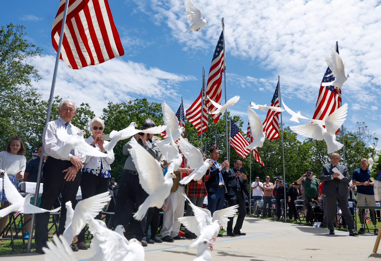 Dozens of people stand to watch a flock of white doves fly into the sky at Oakhill Memorial Park. They are surrounded by American flags and trees during the cloudy but sunny day.
