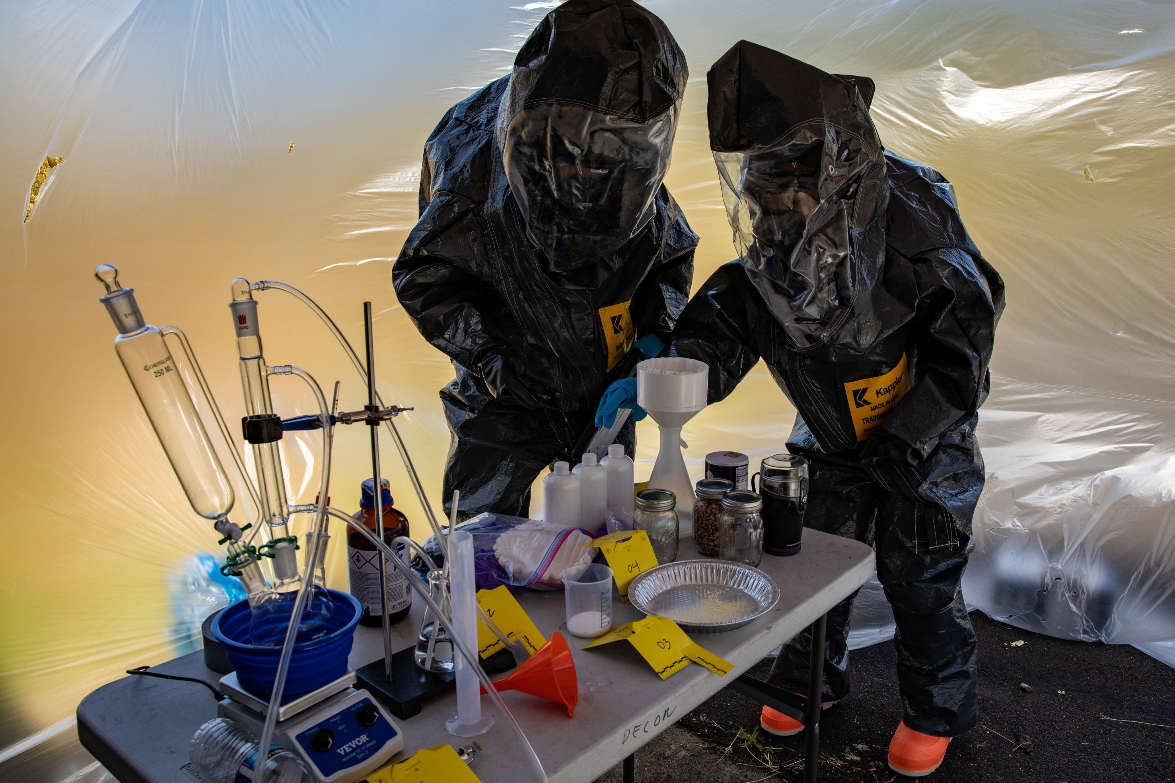 Service members in black HAZMAT suits examine bottles on a table