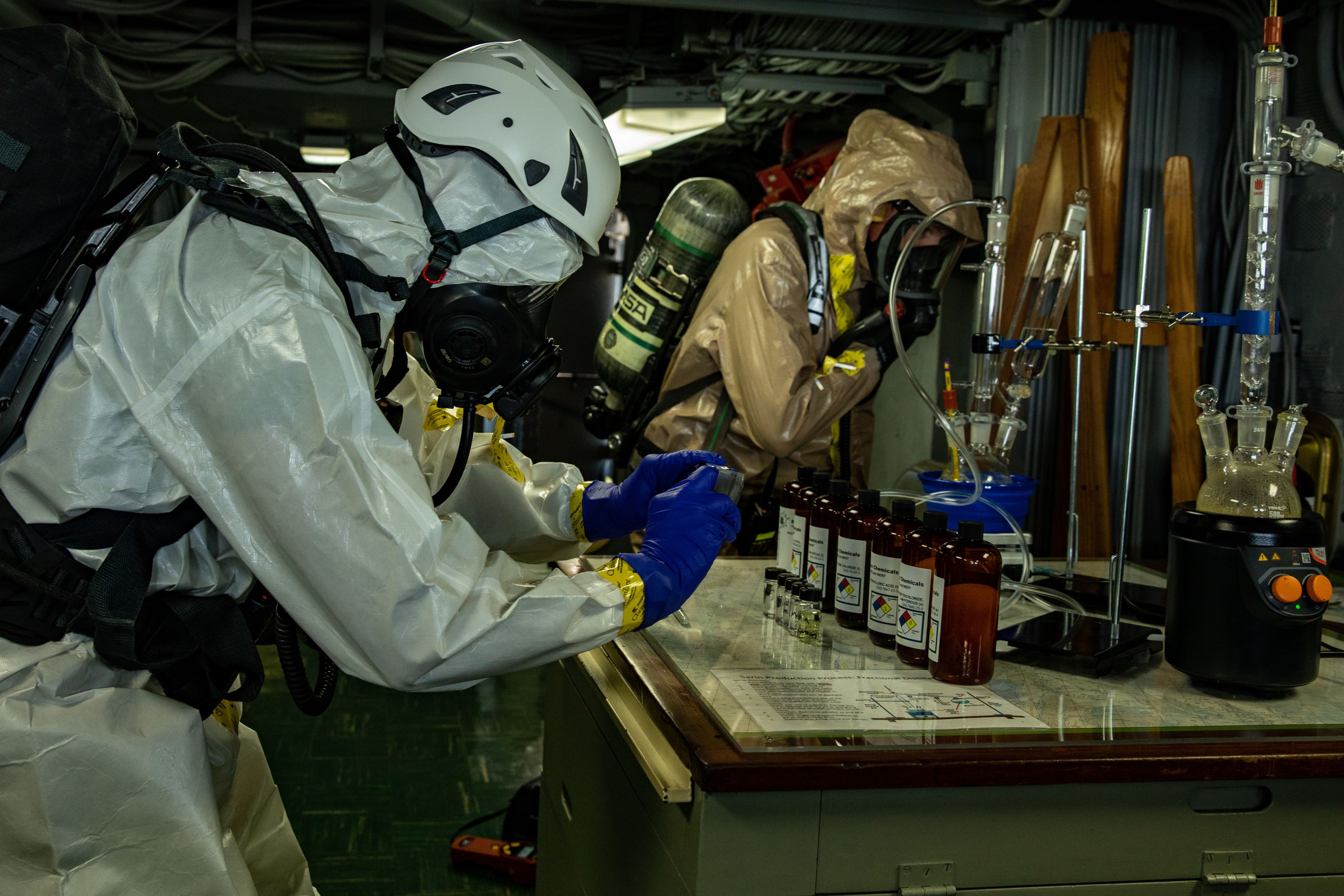 Service members in HAZMAT suits examine bottles and beakers on a table