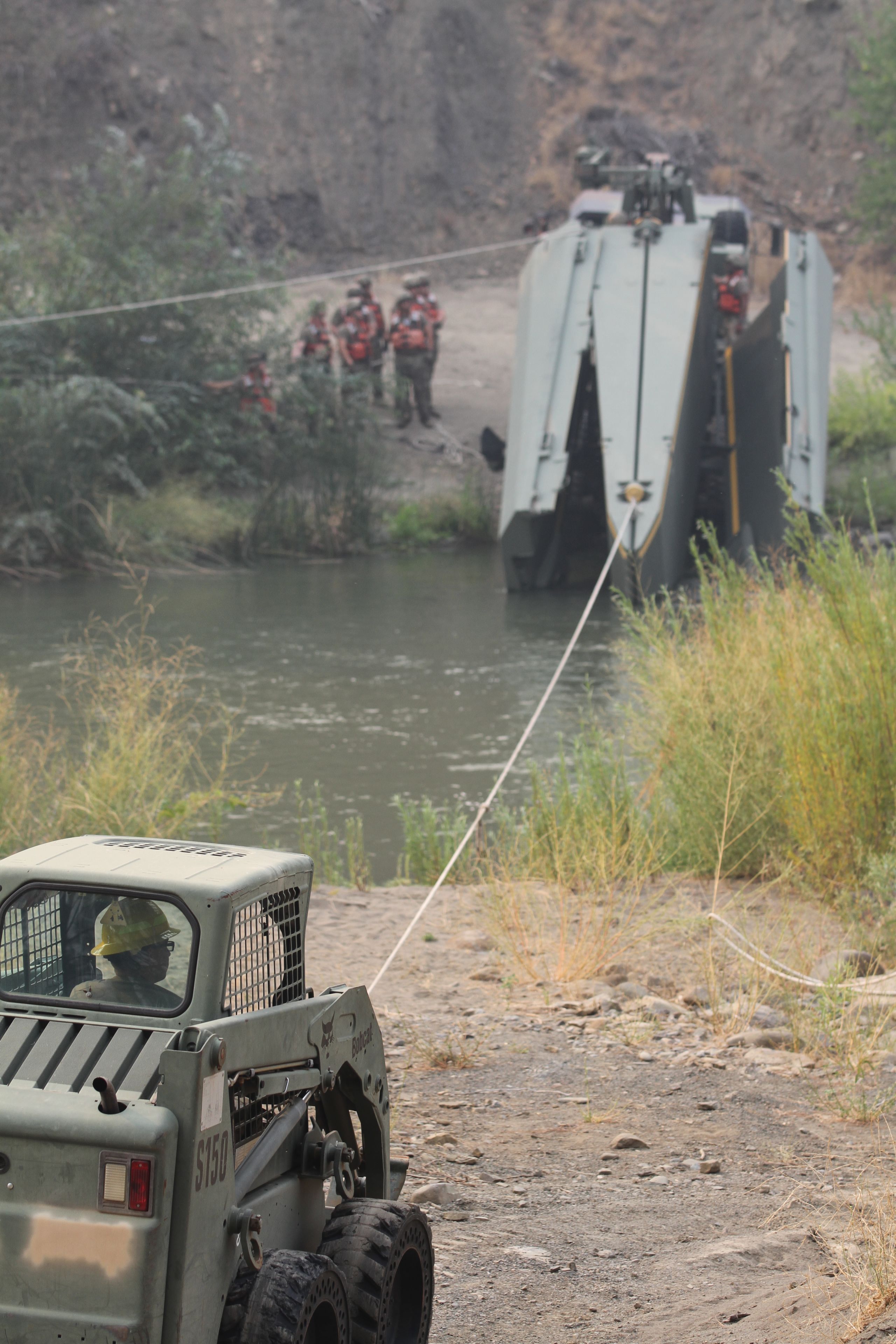 Small tractor pulls floating bridge segment across river by chain.