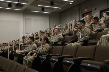 a crowd of soldiers sits in an auditorium and listens to a speaker