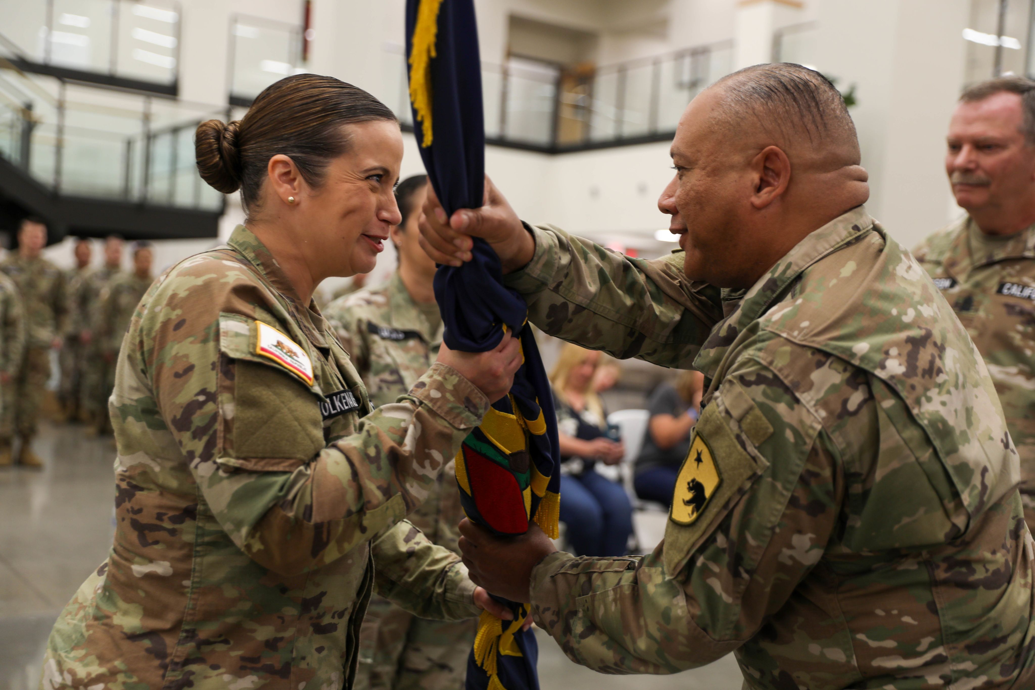 A man in camouflage uniform passes a rolled up flag to a smiling woman in camouflage uniform.