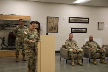 A woman in uniform gives a speech from behind a wooden podium while men in uniform listen