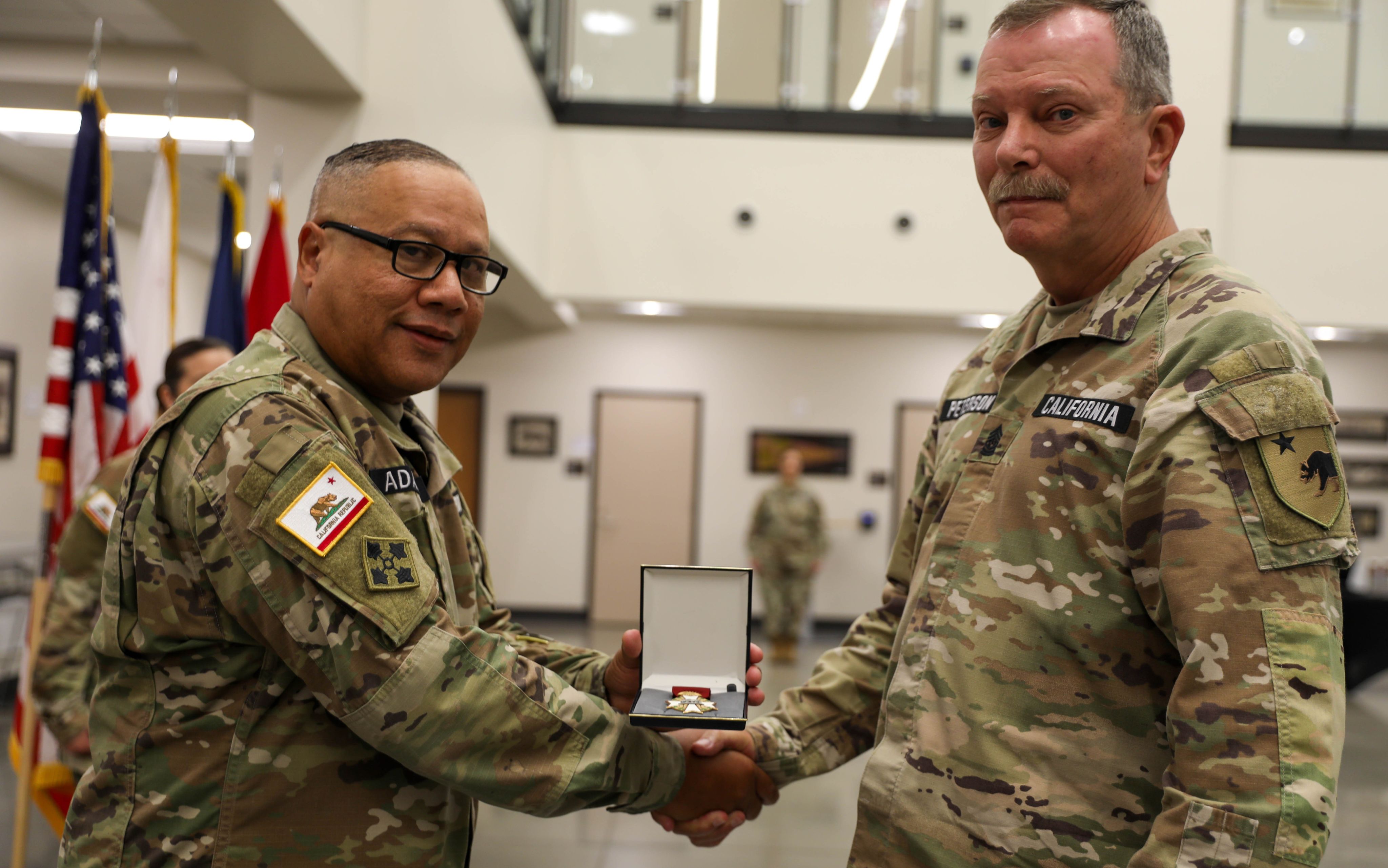 Two men in uniform shake hands as one presents the other with an award.