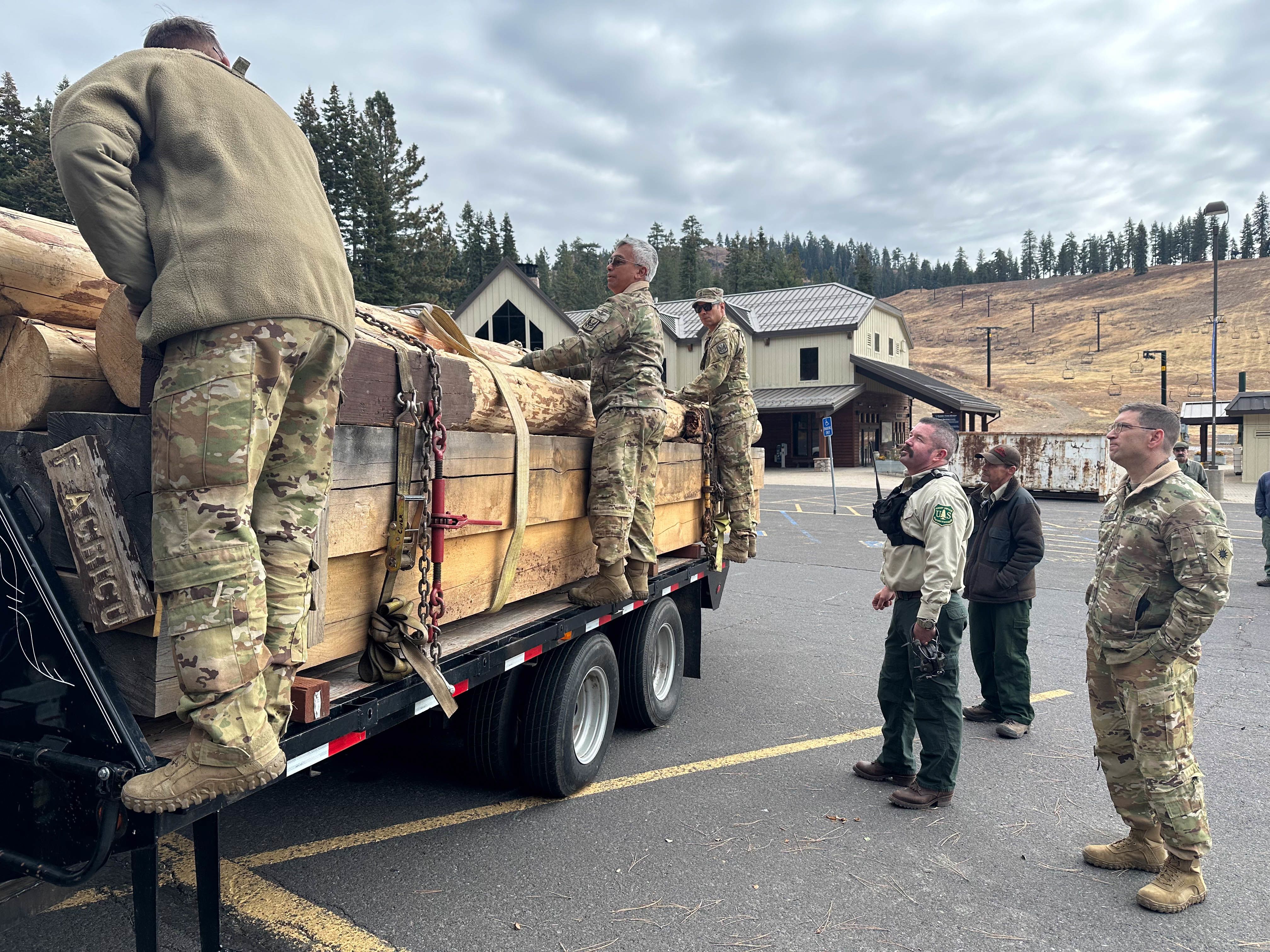 Army Soldiers and Forest Service Employees unload timber logs in a flatbed trailer.