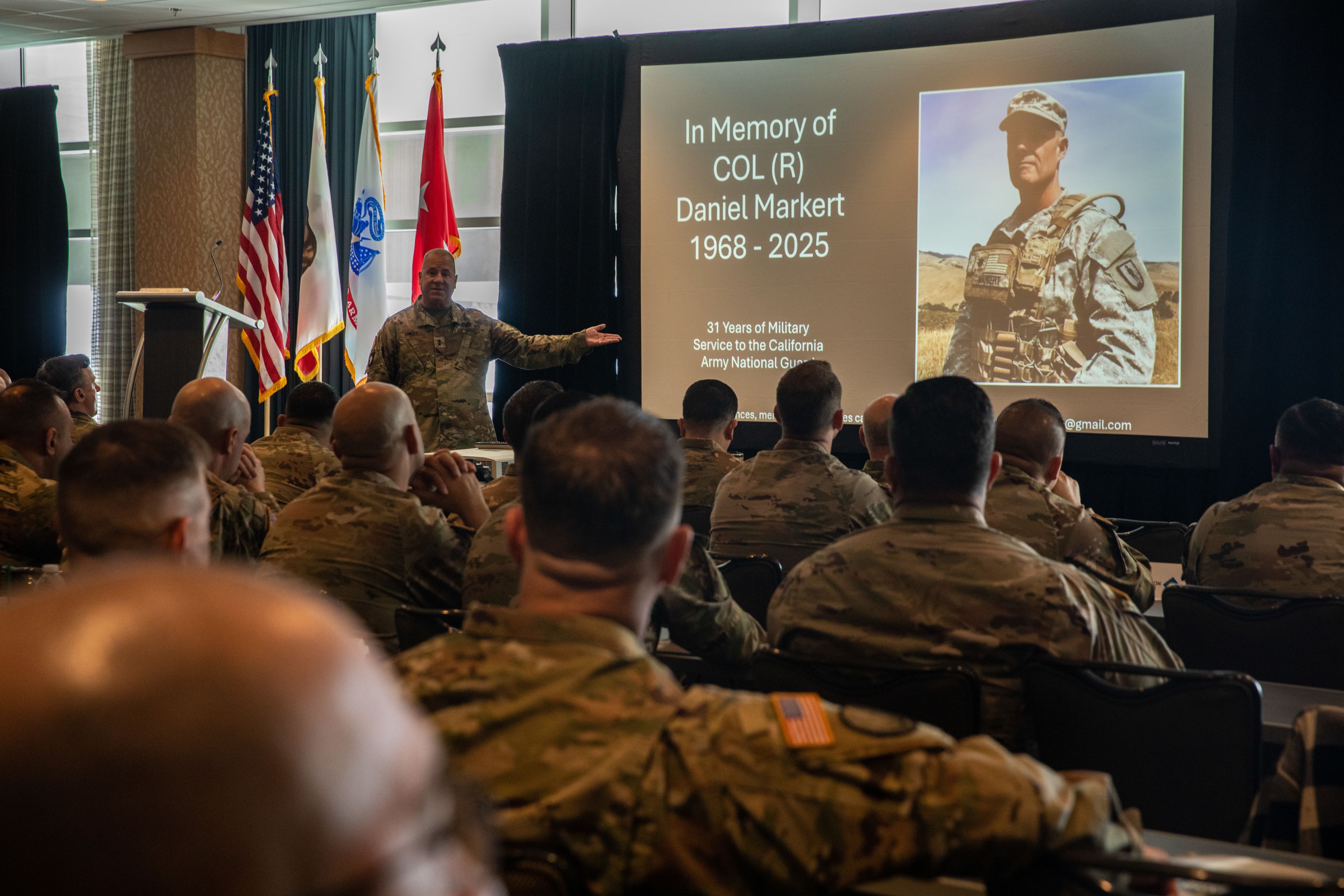 Soldier speaks to an audience while motioning to a screen