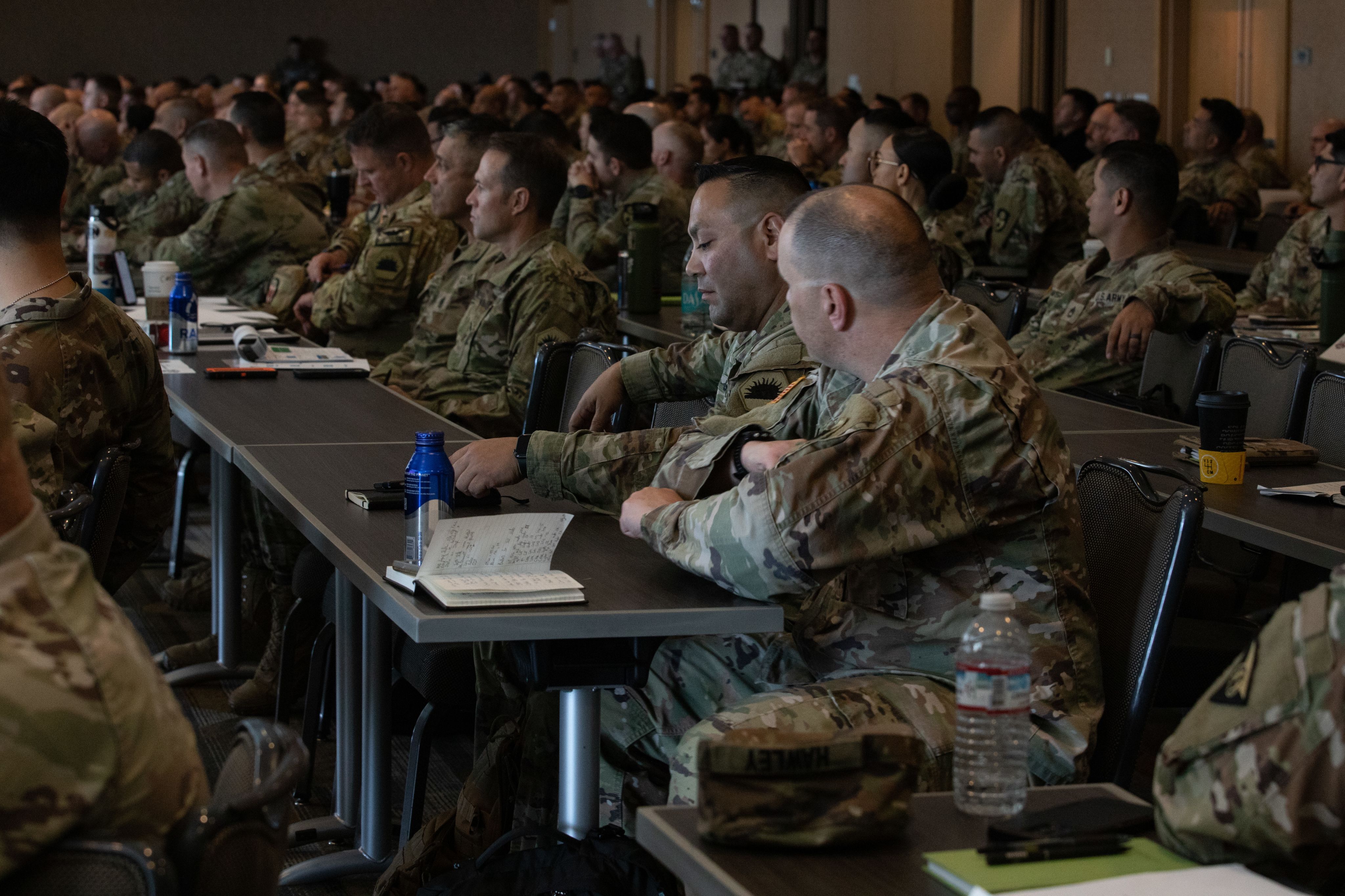 Numerous Soldiers sit at tables while listening to a brief