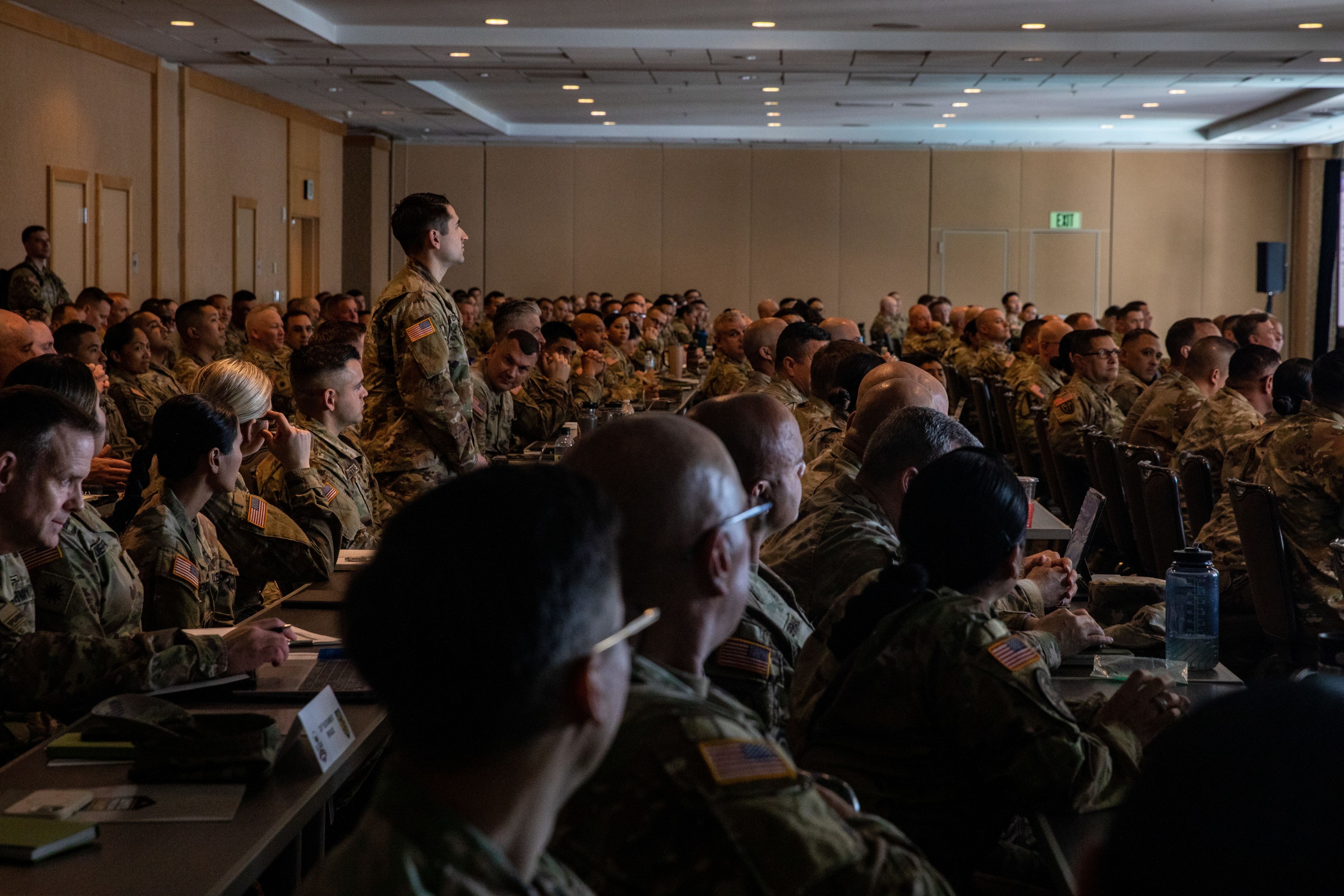Numerous Soldiers sit at tables while listening to a brief
