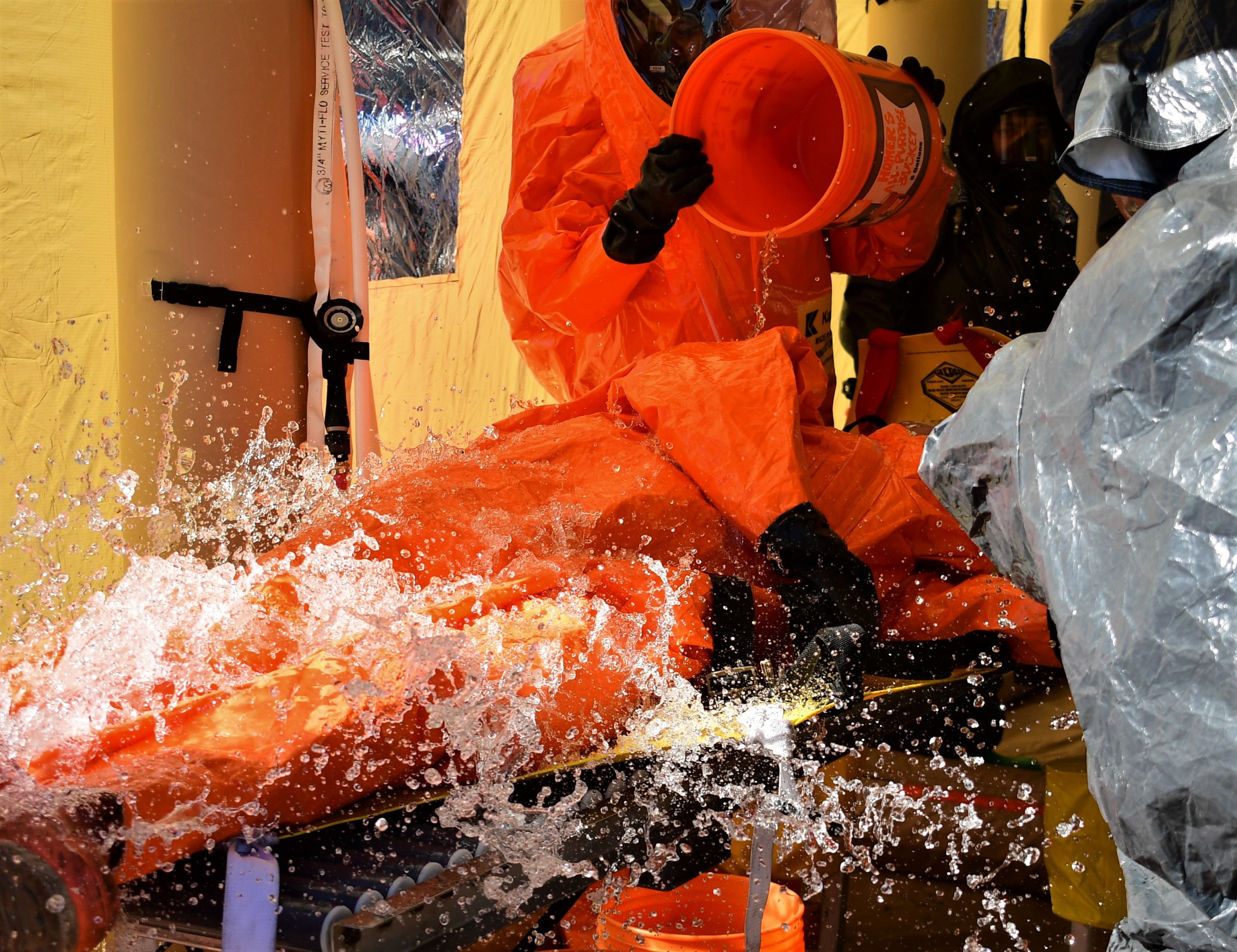 Hazmat clad servicemembers toss buckets of water over a fellow servicemember to decontaminate the suit from hazardous chemicals.