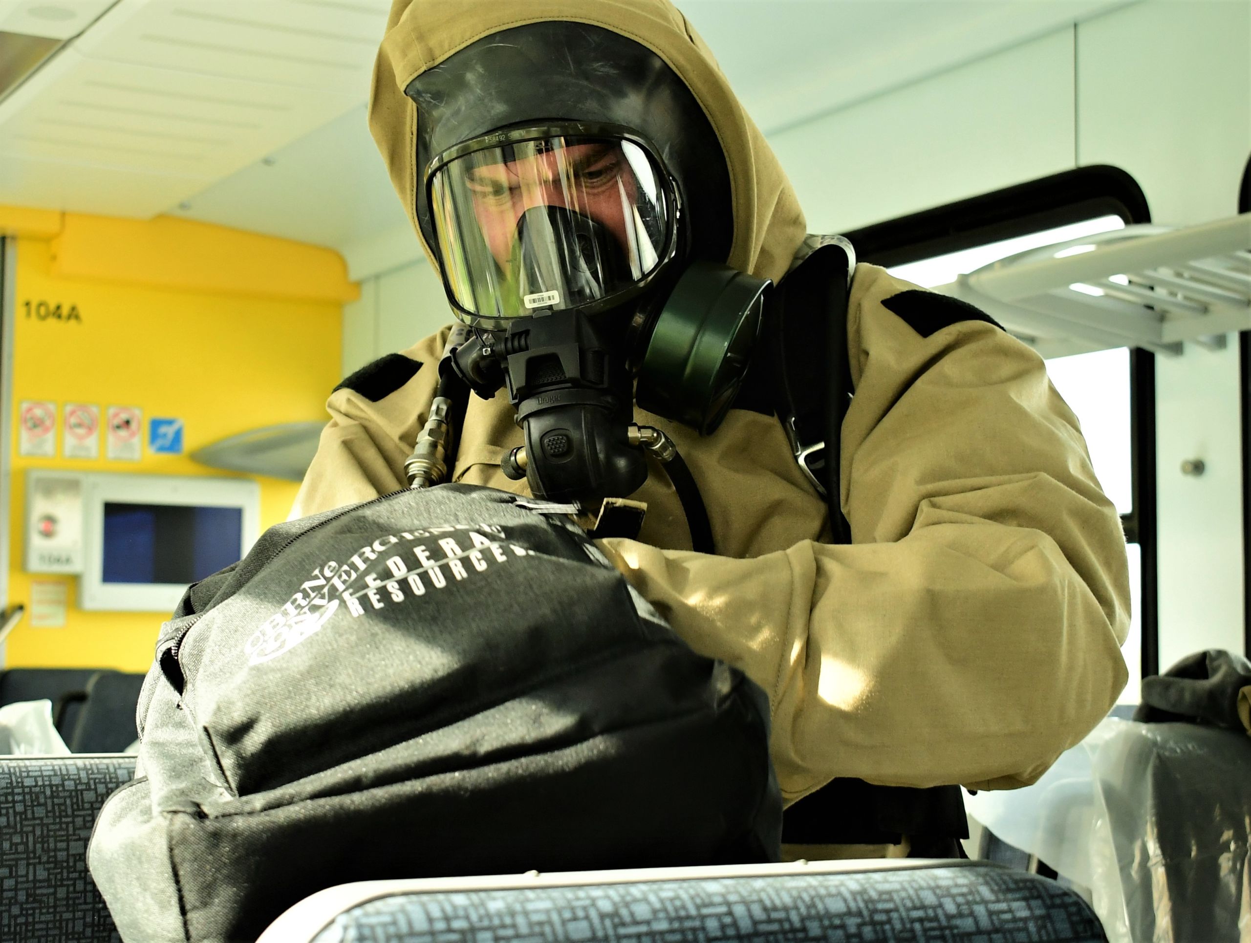 A servicemember in a hazardous material suit and oxygen respirator inspects a black backpack for hazardous materials.