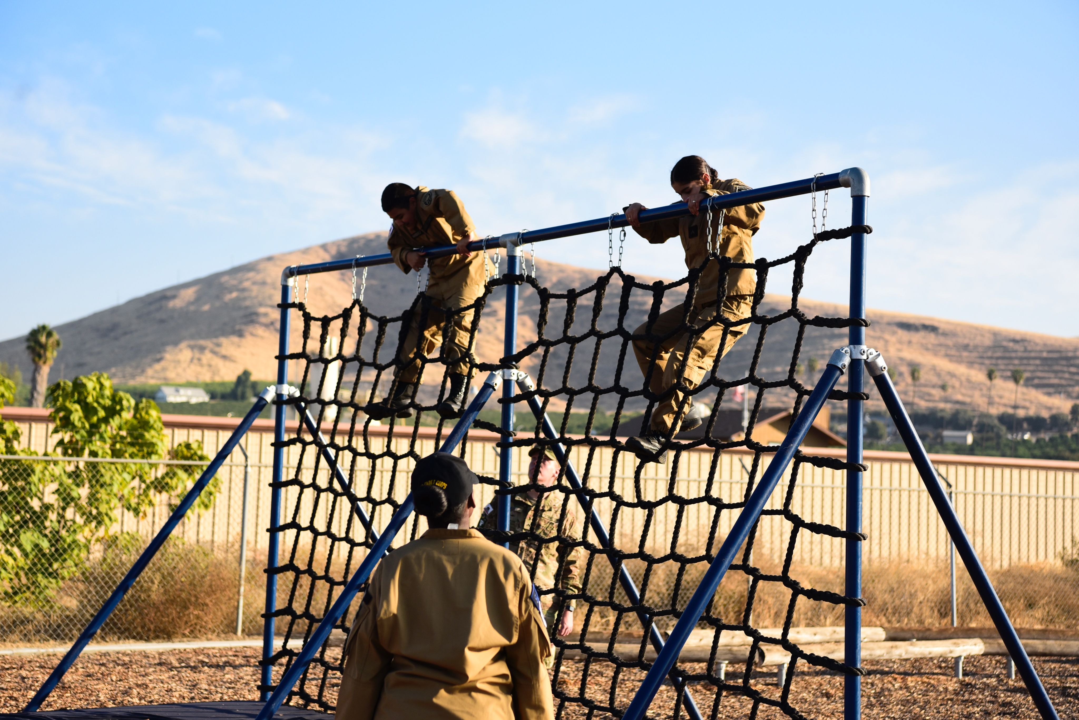 2 cadets climb rope wall while cadets watch them climb.